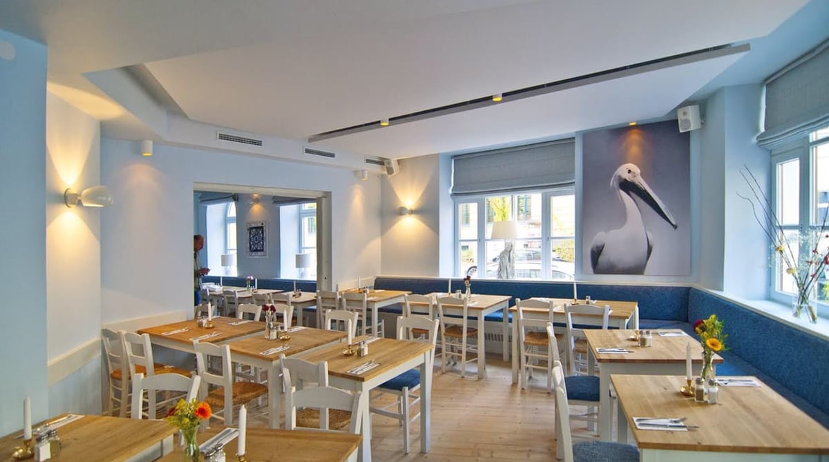 Gastraum der Taverna Melina mit Holztischen, blauen Sitzbänken und einem Pelikan-Motiv an der Wand