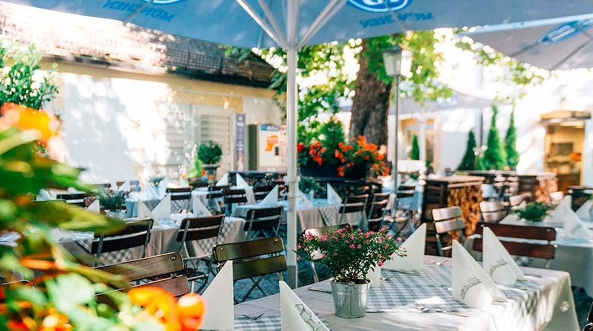 Gedeckte Biergartentische mit Blumen unter Sonnenschirmen vor dem Schwaige-Gebäude