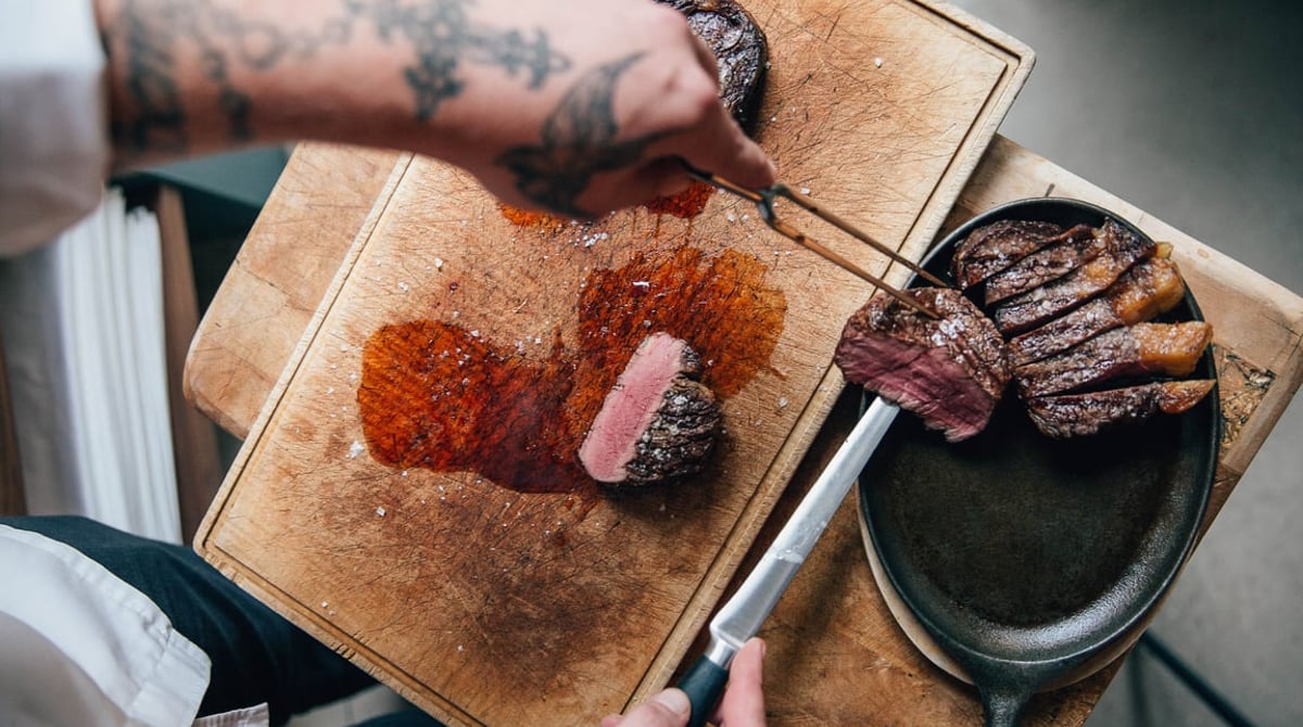 Gegrilltes Steak wird auf einem Holzbrett aufgeschnitten, daneben eine gusseiserne Pfanne
