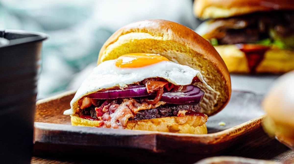 Burger mit Spiegelei, Bacon und roten Zwiebeln im Brioche-Bun auf einem Holzbrett