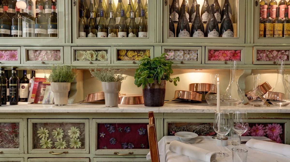 Gedeckter Tisch vor dem mintgrünen Schrank mit Weinflaschen, Blumenfächern und Kupfertöpfen im Vinaiolo