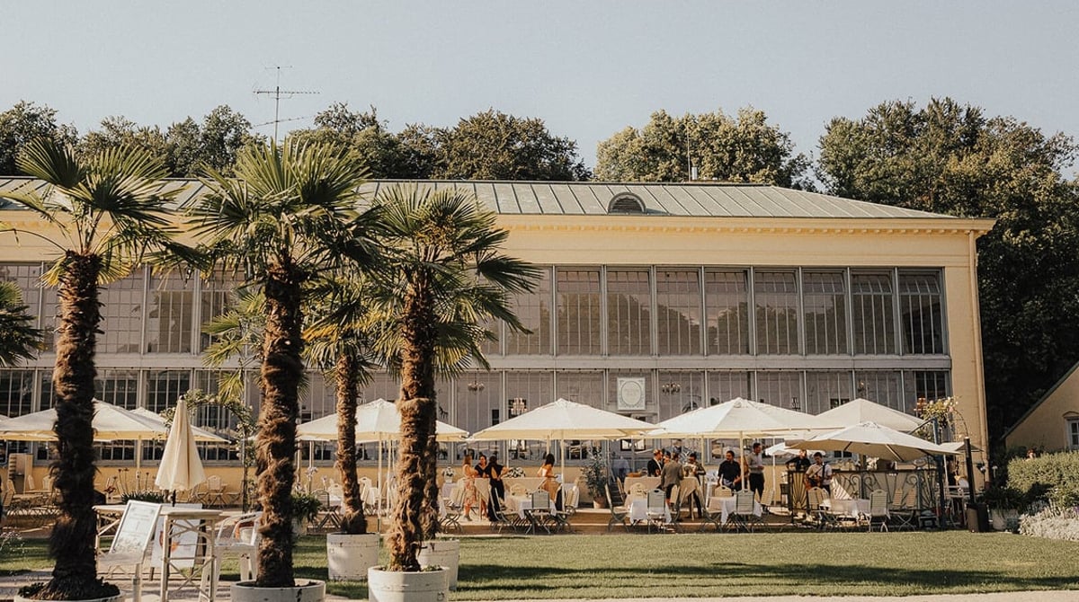 Der Glasbau des Palmenhauses im Schlosspark Nymphenburg mit Palmen und Sonnenschirmen auf der Terrasse davor