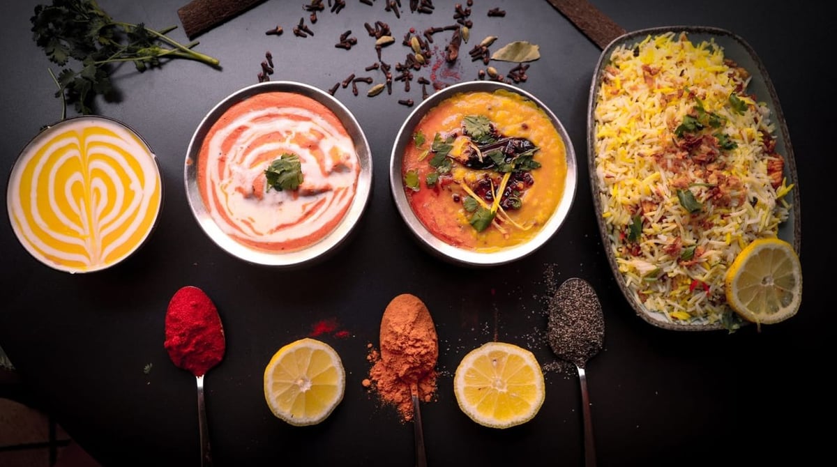 Vier Currys in Schalen und Biryani auf dunklem Grund, davor Gewürze auf Löffeln