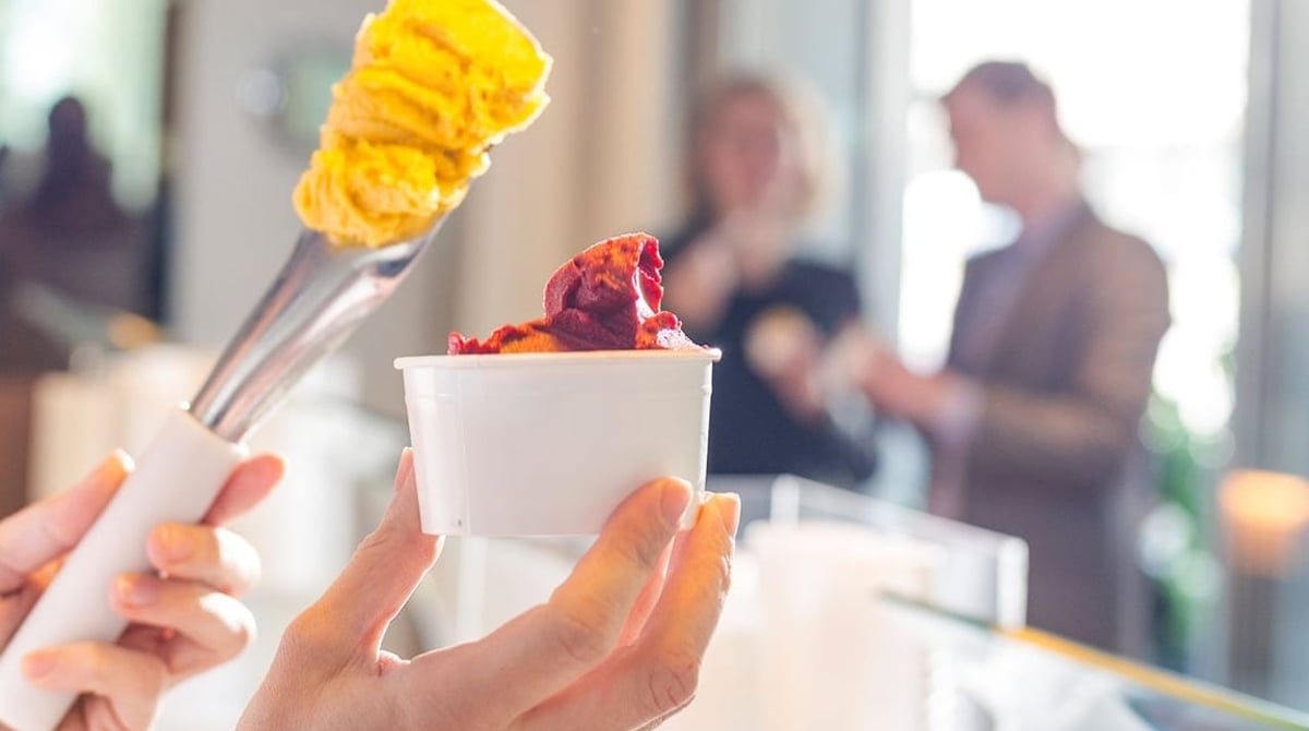 Becher mit gelbem und rotem Eis, in der Hand gehalten vor der Theke