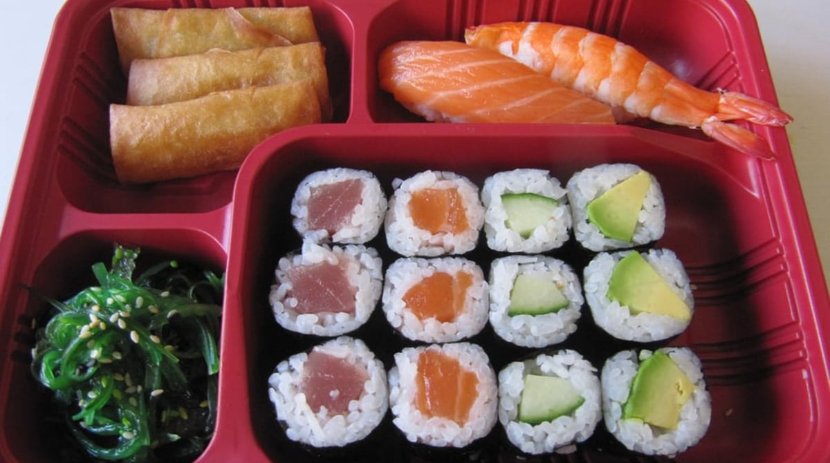 Bento-Box in roter Schale mit Frühlingsrollen, Lachs- und Garnelen-Nigiri, Maki mit Thunfisch, Lachs, Gurke und Avocado sowie Algensalat