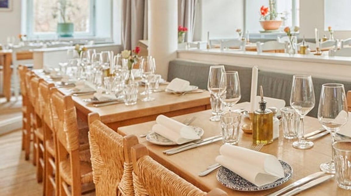 Langer Gastraum der Taverna Paros mit eingedeckten Holztischen und Korbstühlen