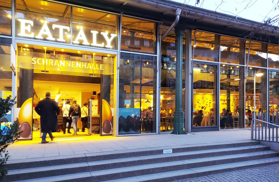 Eingang von Eataly in der Schrannenhalle am Viktualienmarkt mit leuchtendem Schriftzug und Glasfassade in der Abenddämmerung