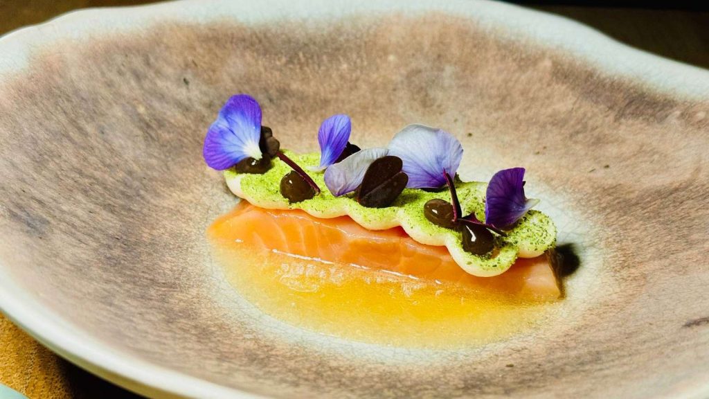 Gabelspiel - IN München - Das Stadtmagazin Salmon fillet topped with a green herb crumble, dark sauce dots, and purple edible flowers on a shallow plate.