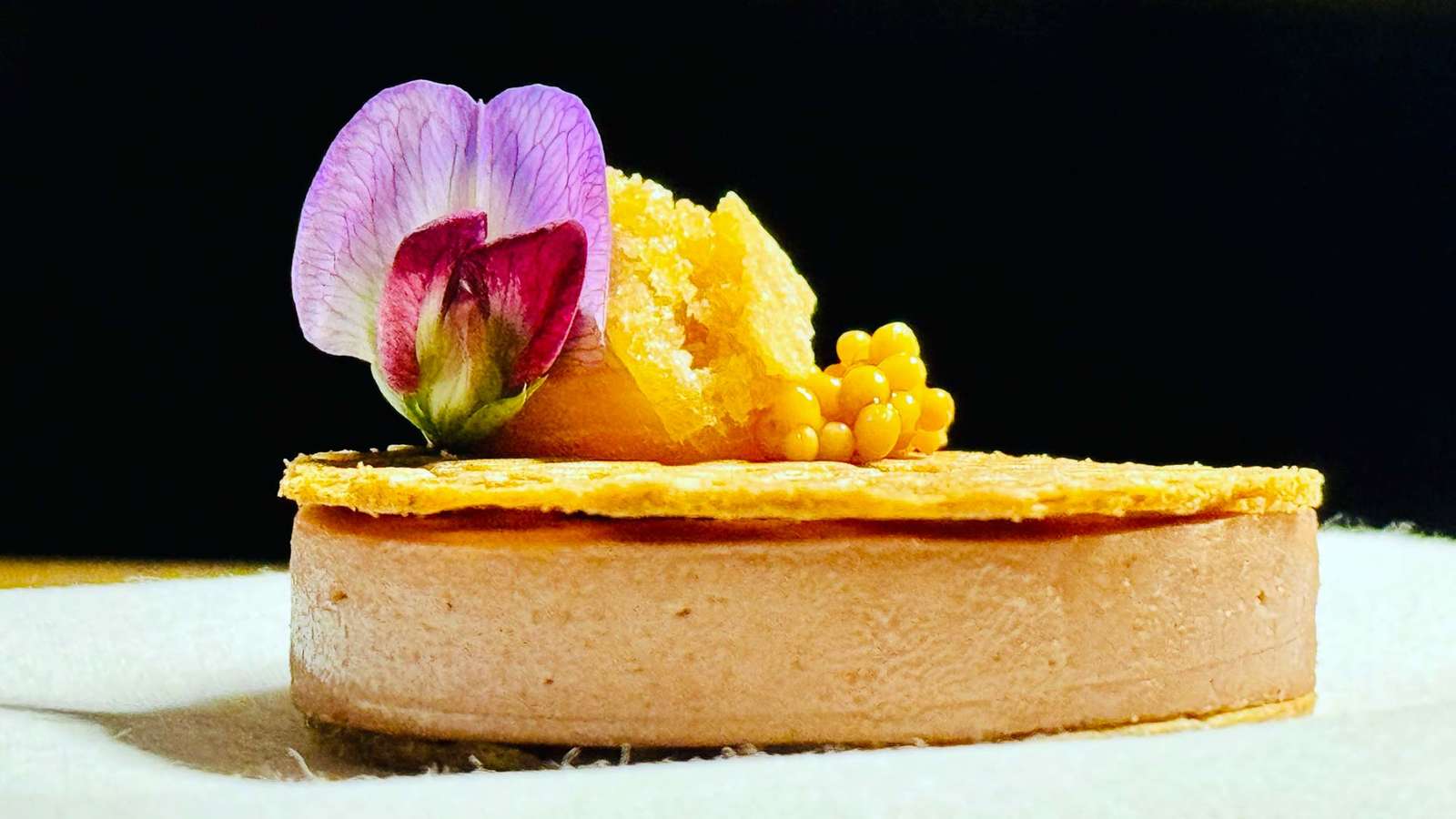 Rundes Gericht auf knusprigem Teig mit Blüte, orangefarbener Creme und gelben Perlen vor dunklem Hintergrund