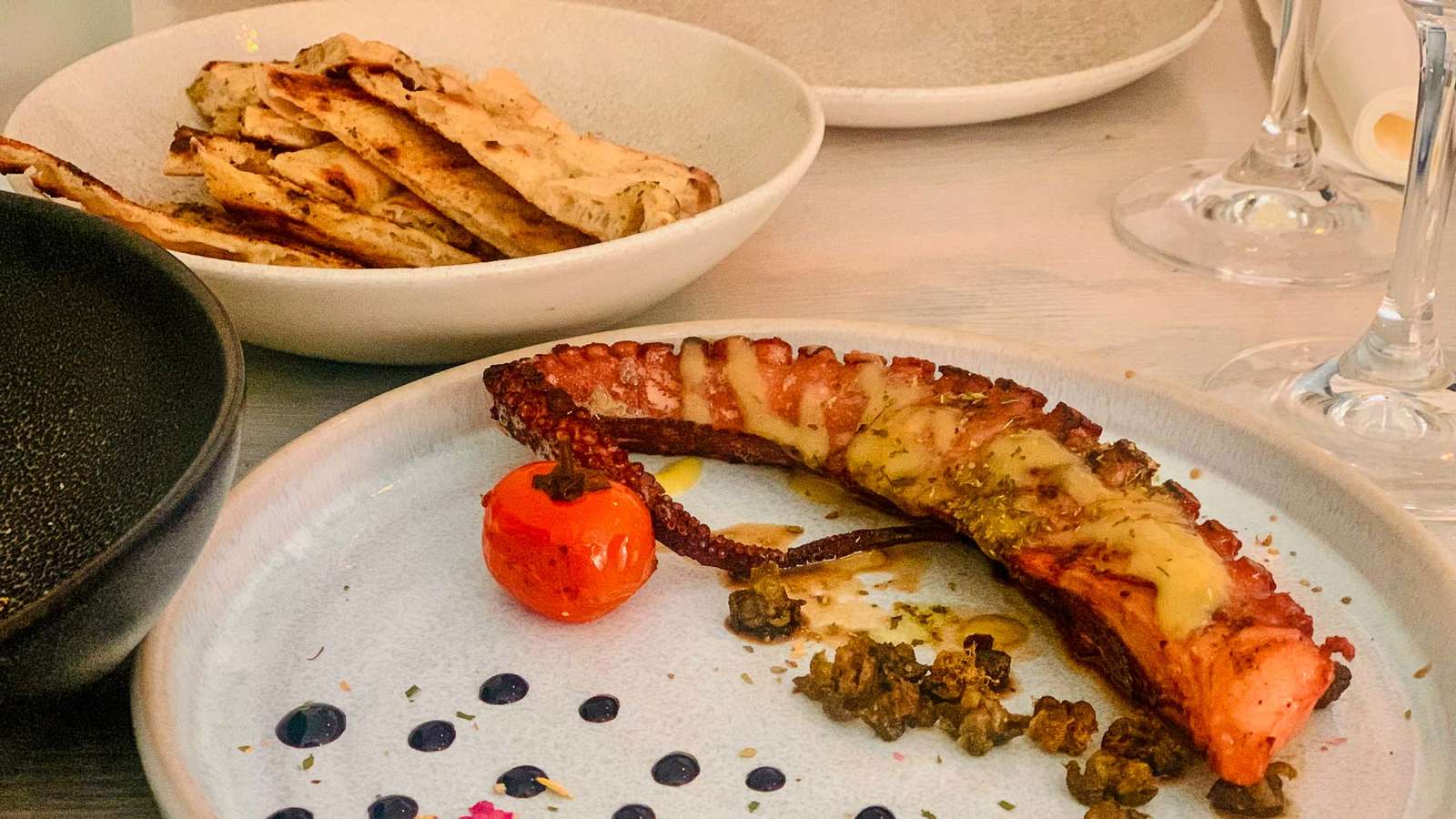 Gegrillter Oktopusarm mit Kapern und Kirschtomate auf einem hellen Teller, daneben Fladenbrot