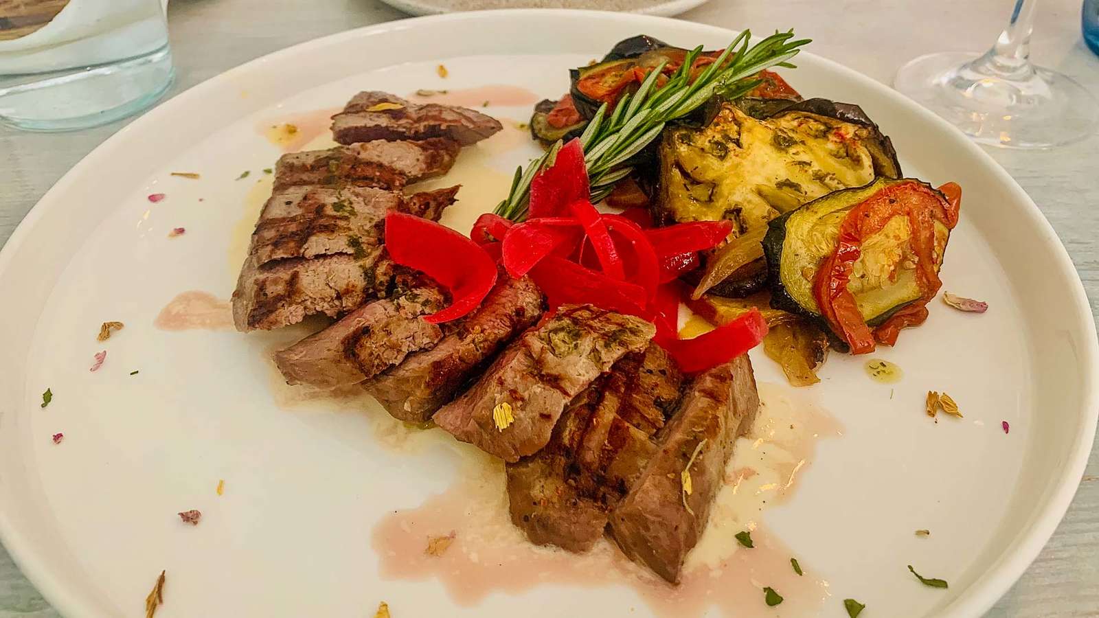 Gegrillte Lammlachse mit geschmortem Gemüse, roten Paprikastreifen und Rosmarinzweig auf einem weißen Teller