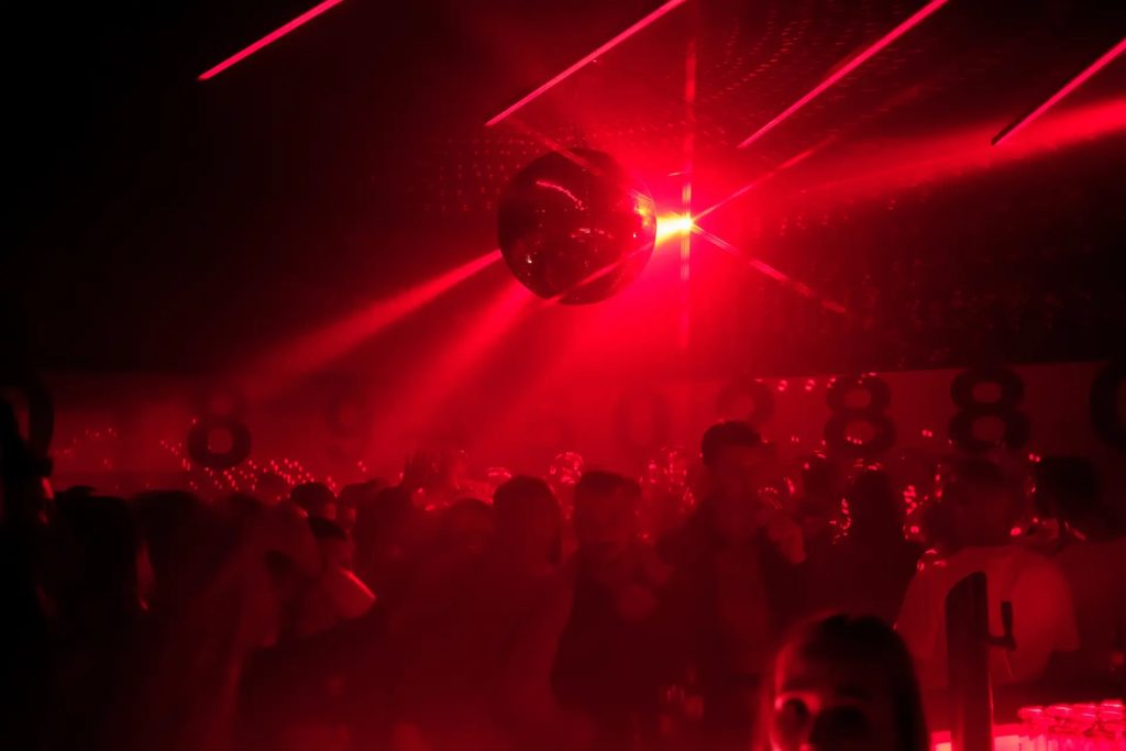 Red-lit nightclub scene with a suspended disco ball and a dense crowd dancing beneath bright beams of light.