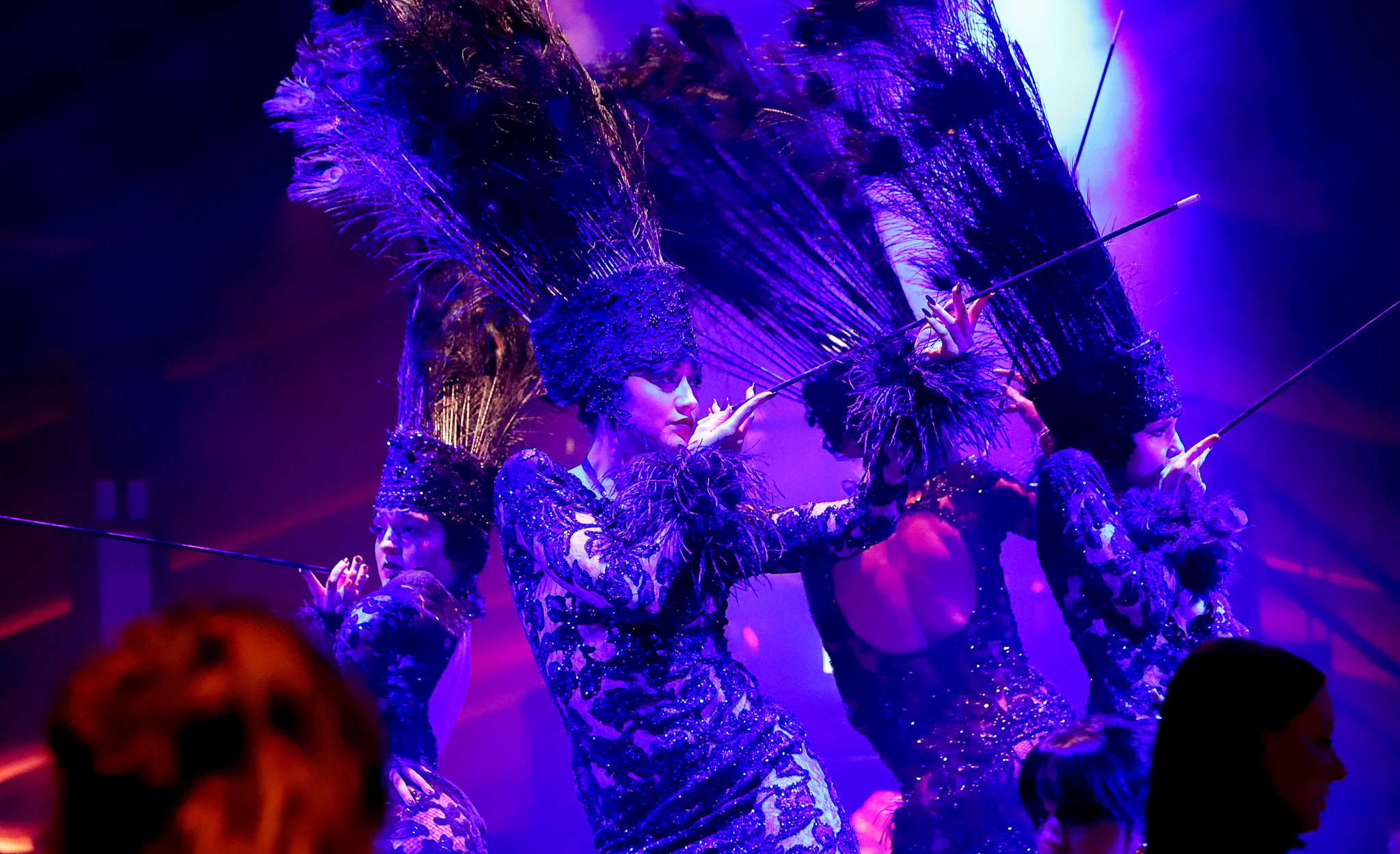 Group of dancers in sequined purple costumes and tall feathered headdresses performing on a purple-lit stage.