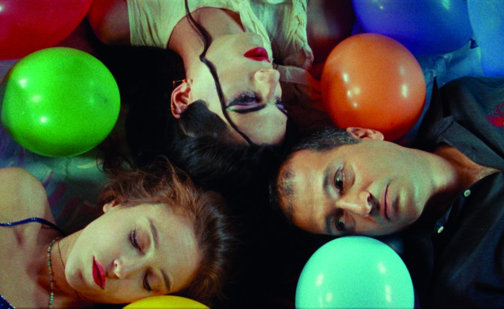 REIHEN UND FESTIVALS 10_26 2 - IN München - Das Stadtmagazin Four friends lying in a circle with colorful balloons around them at a party, heads close together and eyes closed or relaxed.