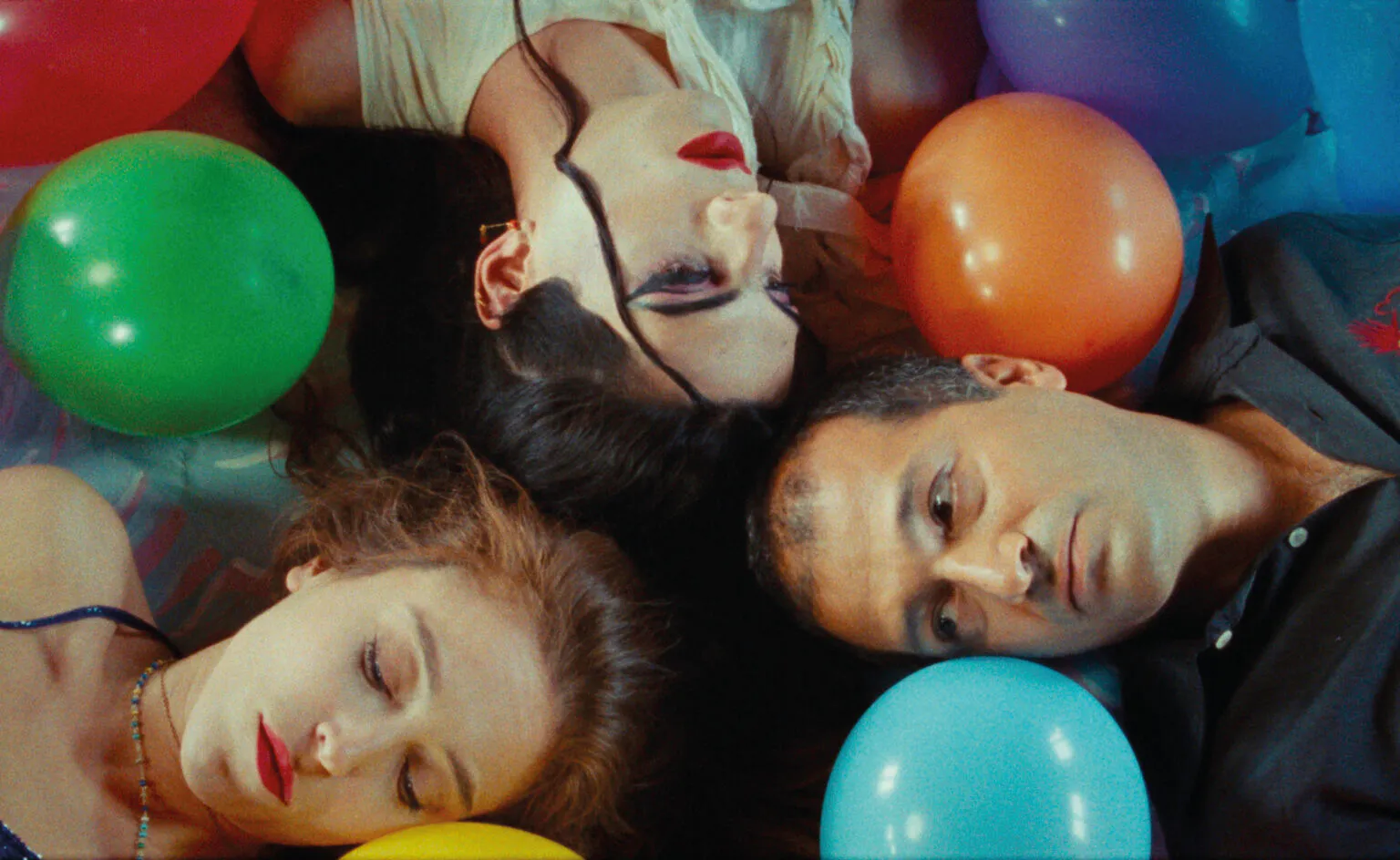 Four friends lying in a circle with colorful balloons around them at a party, heads close together and eyes closed or relaxed.