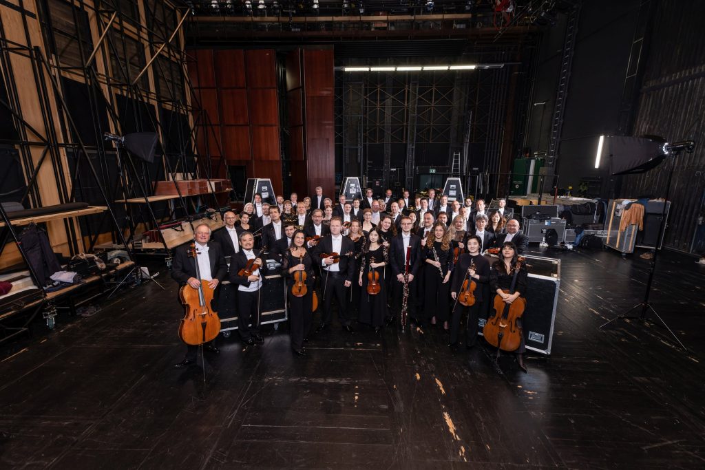 Das Münchner Rundfunkorchester - IN München - Das Stadtmagazin Large orchestra group in formal wear posing for a portrait on backstage theater stage with instruments visible (violas, violins, cellos) and stage equipment around them.