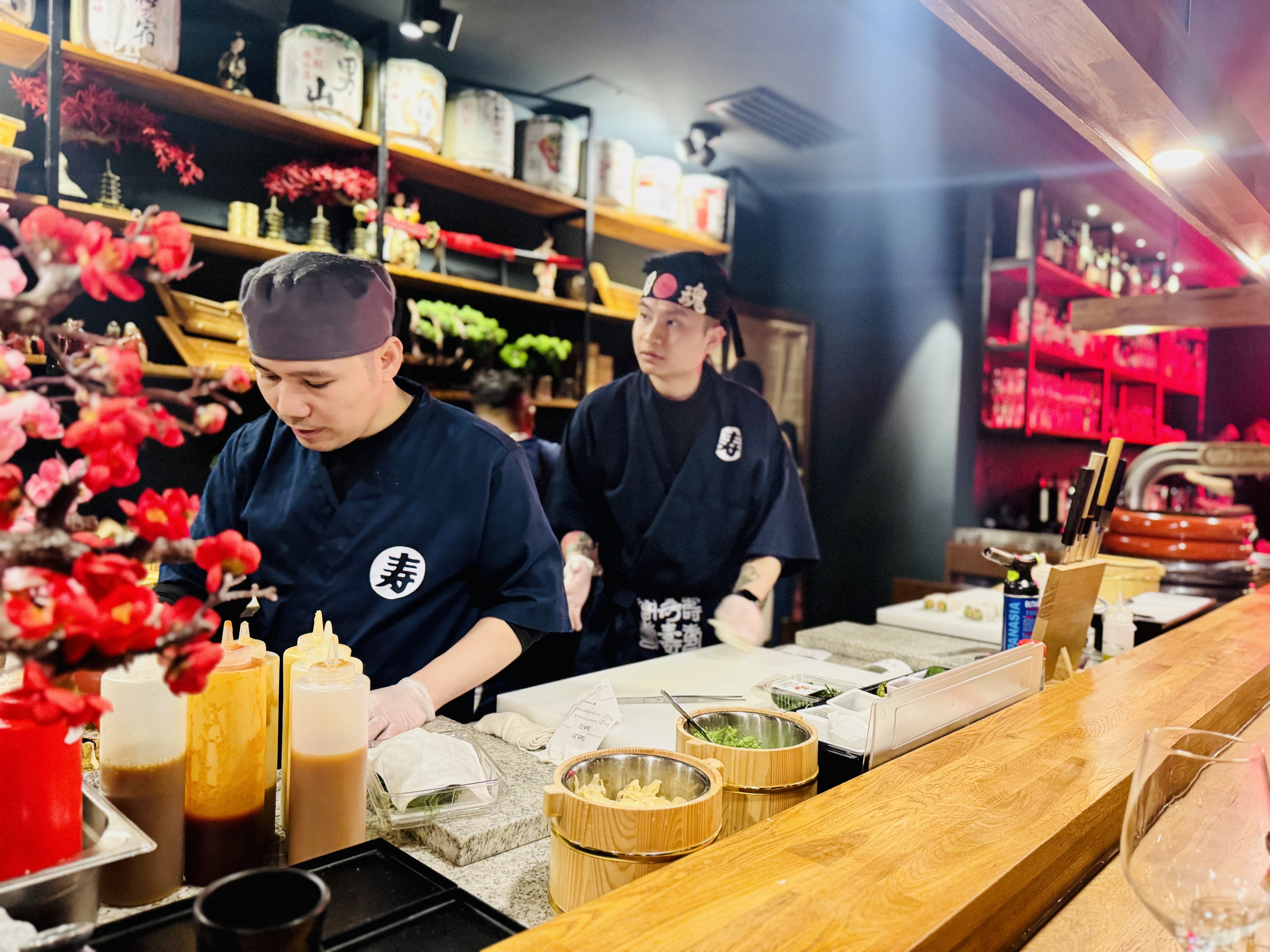Zwei Sushi-Köche in dunkelblauen Jacken bereiten hinter der Theke Speisen zu, davor rote Blüten und Saucenflaschen