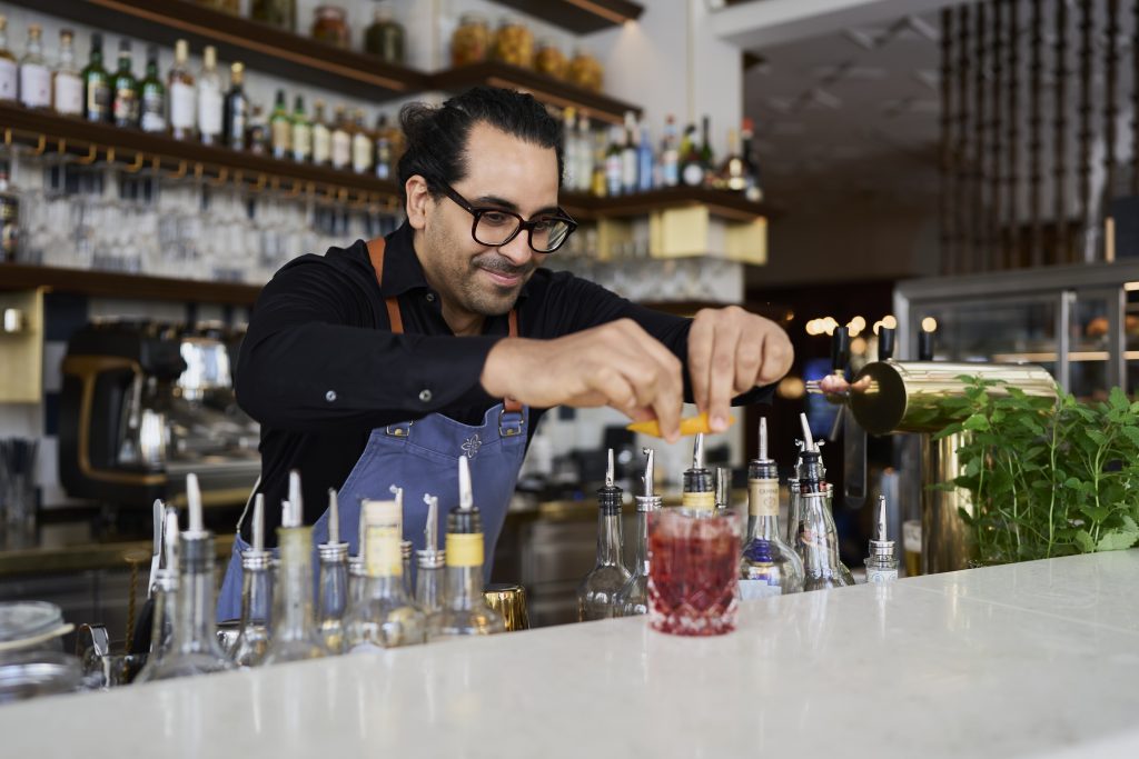 Josefa Team - IN München - Das Stadtmagazin Bartender wearing glasses and a blue apron prepares a drink at a bar, with bottles and a red cocktail on the counter behind him.