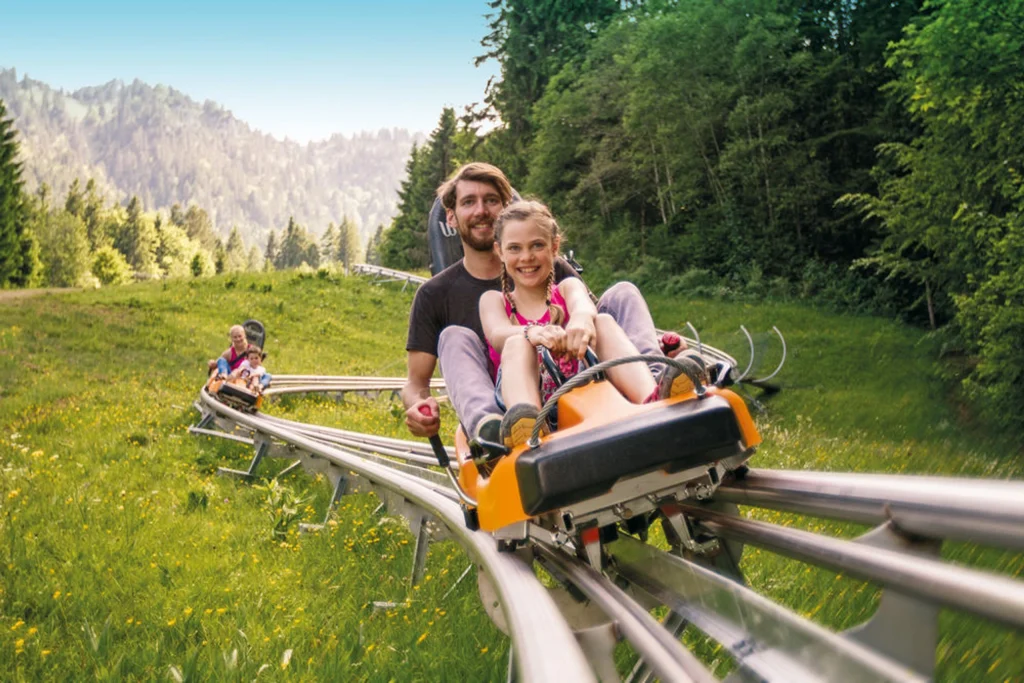 Kolbensattel-Alpine-Coaster-3 - IN München - Das Stadtmagazin A smiling man and girl ride a roller coaster along a metal track through a green meadow with trees and distant mountains in the background.