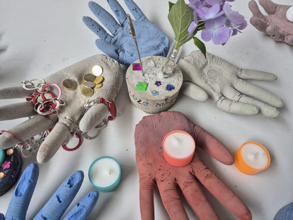 Brücken bauen 20250806_153540 - IN München - Das Stadtmagazin Multiple colored clay hands (blue, pink, gray) arranged on a table among jewelry, coins, and small craft items, with a vase of purple flowers nearby.