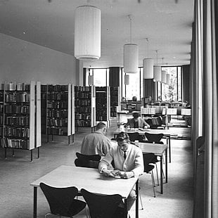Schwarz-weiß-Foto von Personen die im Recherchezentrum (ehemalige Bibliothek) des Amerikahaus an Tischen lesen. Daneben sind Bücherregale aufgereiht ©Amerikahaus München