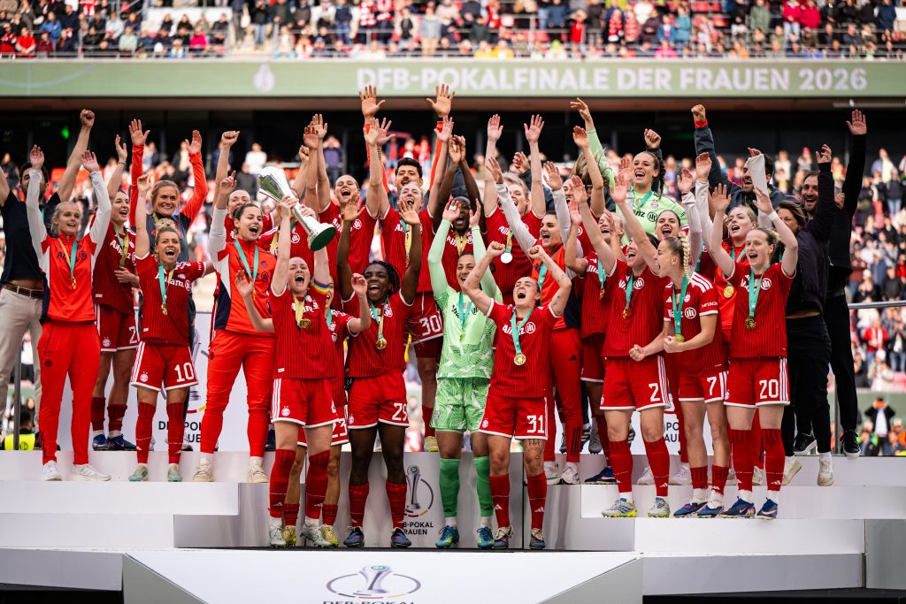 Female soccer team on a podium celebrate with trophy and medals, raising arms amidst cheering crowd.