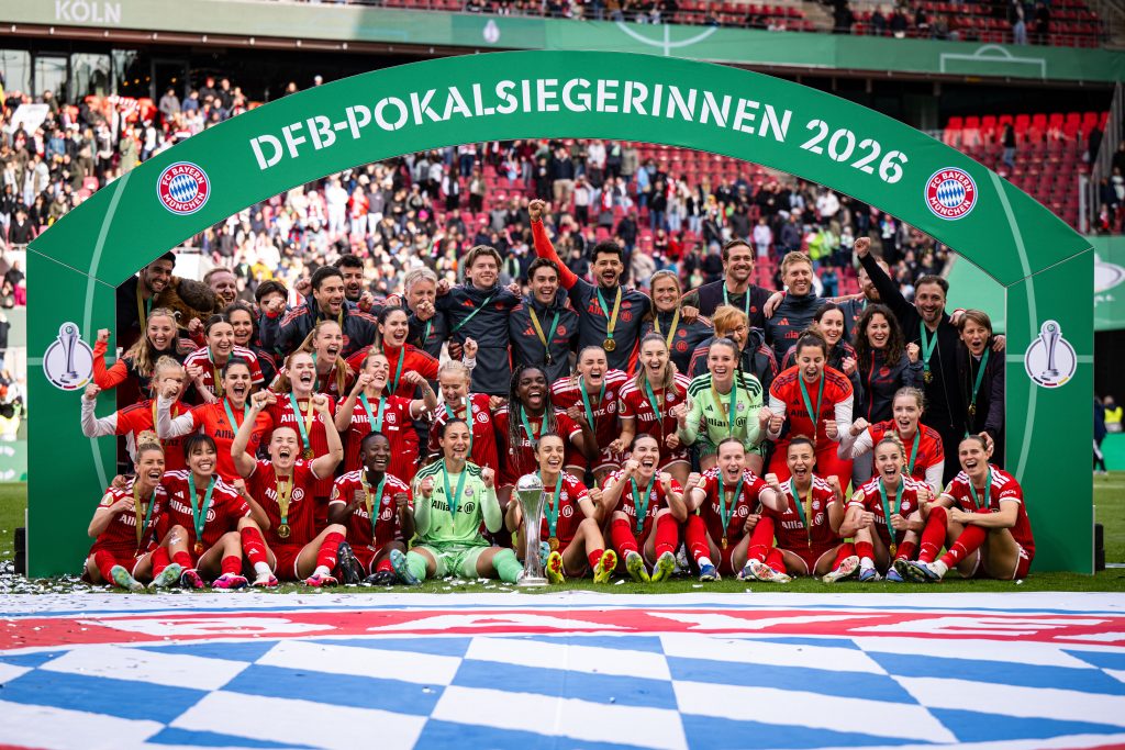 FC Bayern women’s football team pose with medals and trophy under a green arch reading 'DFB-POKALSIEGERINNEN 2026' after winning the cup. Fans cheer in the stands behind them.