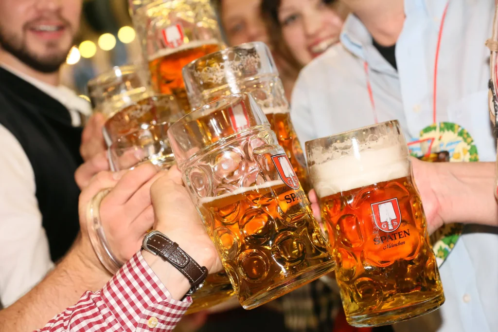 Im Wiesnfestzelt Ochsenbraterei © München Tourismus Tommy Loesch - IN München - Das Stadtmagazin Friends clinking large beer steins in a toast at a festive gathering; Spaten München branding visible on glasses.