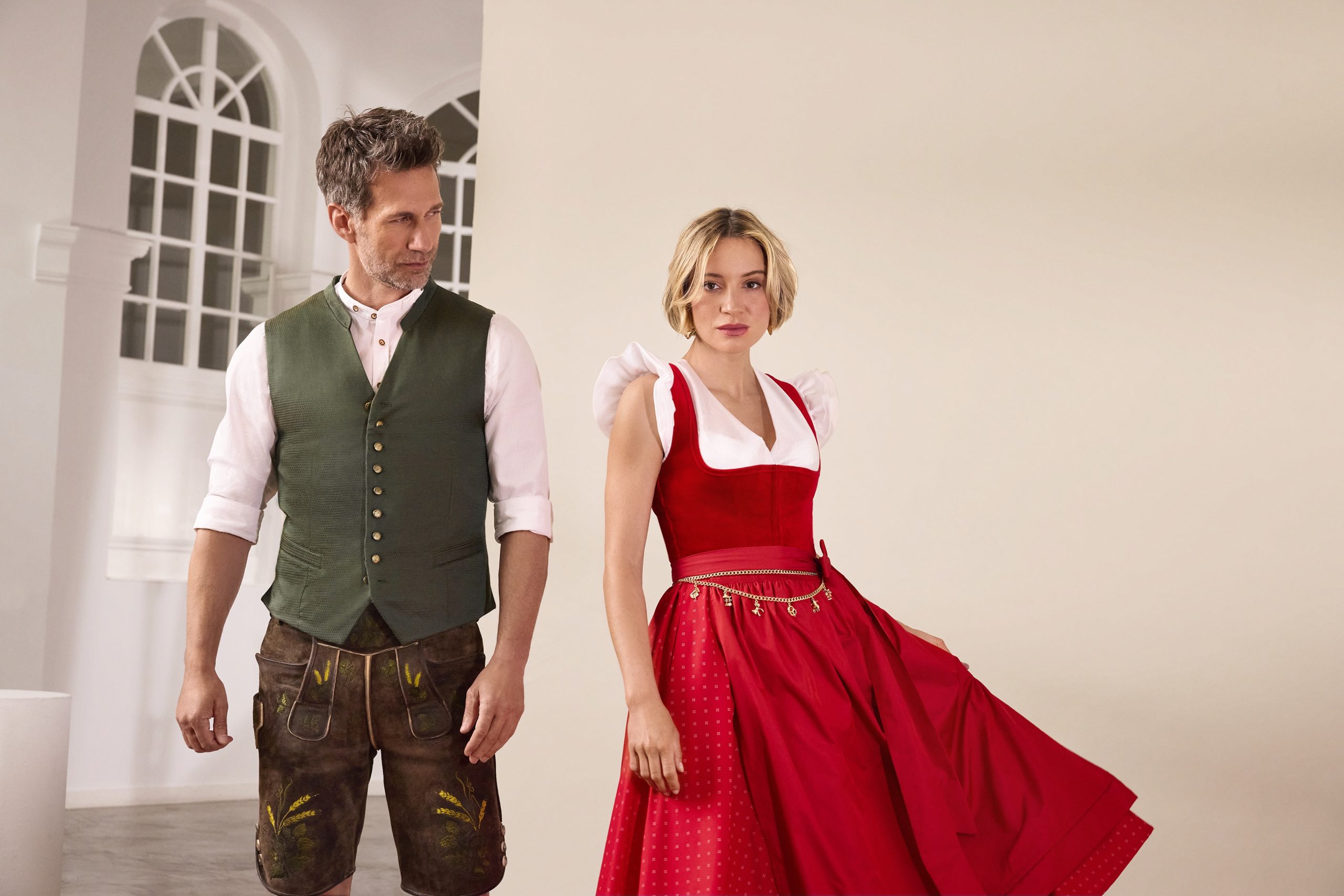 Man in a green vest and brown lederhosen stands beside a woman in a red dirndl indoors.
