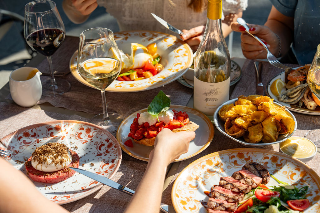 People dining outdoors with a variety of dishes, wine glasses, and a bottle on a patterned tablecloth.