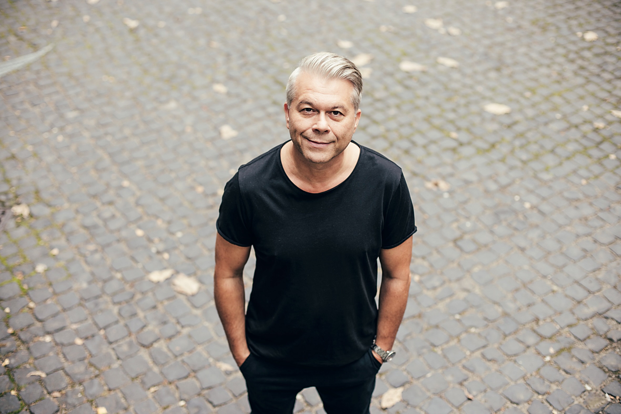 Man with gray hair wearing a black t-shirt stands outdoors on a cobblestone street, looking up at the camera.