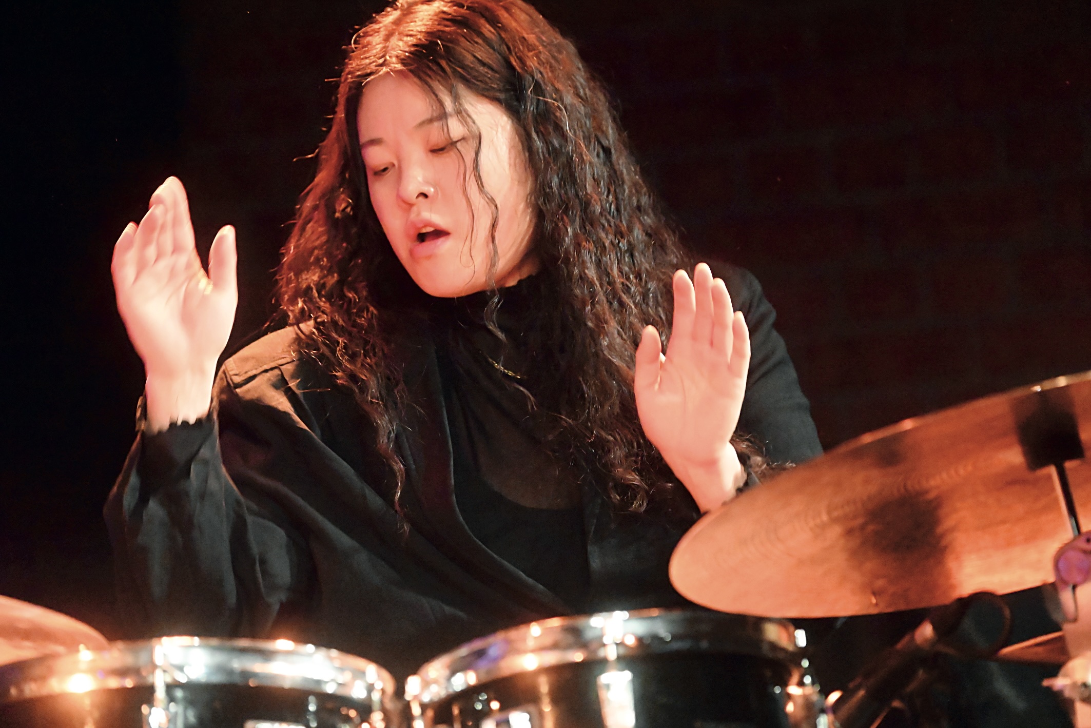 Drummer with long curly hair raises hands mid-performance, playing a drum set in a dark stage.