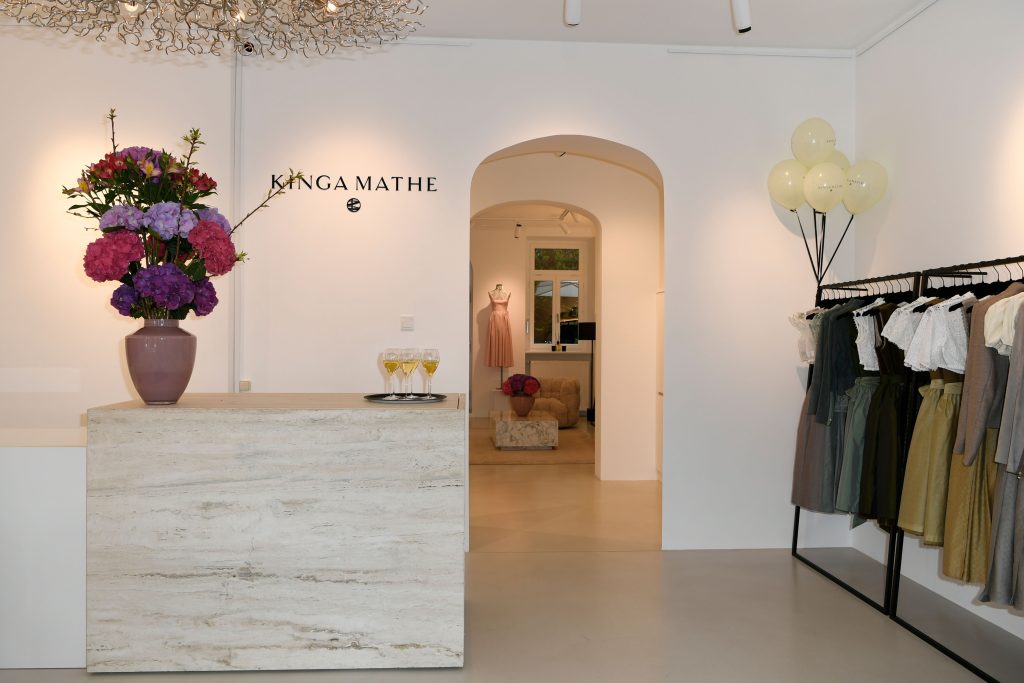 KINGA MATHE Store-Eröffnung in München am 07.05.2026 - IN München - Das Stadtmagazin Boutique reception area with a marble counter, a tall pink vase of pink and purple flowers, and the wall sign 'KINGA MATHE' beside an arched doorway to fitting rooms. Balloons float in the corner and clothing racks line the right side.