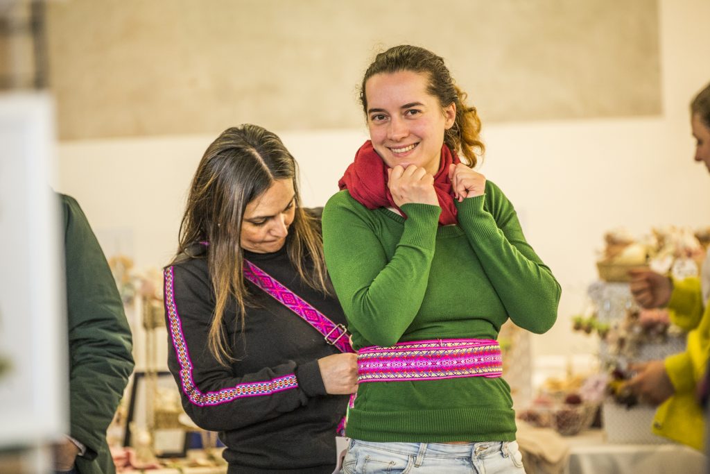 Two women in a shop smile at the camera; one wears a green sweater and red scarf, the other in black with a pink patterned strap across her chest.