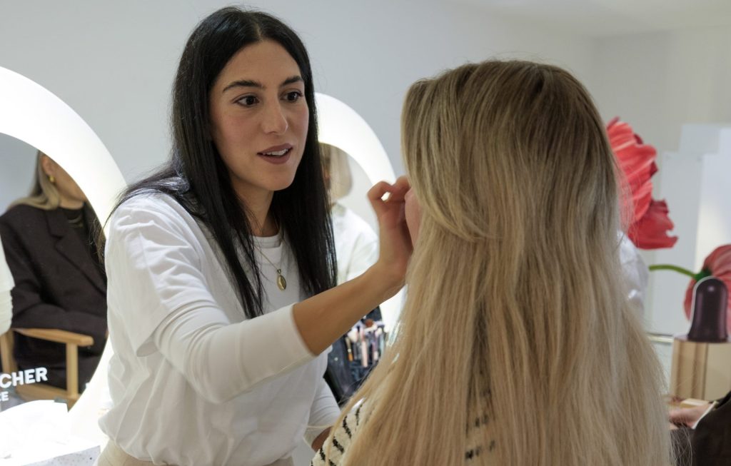 Beauty professional applies makeup to a seated client in a bright studio, with a round illuminated mirror behind them.