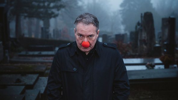 Man in a dark coat with a red clown nose stands in a foggy graveyard.