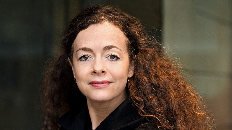 Portrait of a woman with curly brown hair, wearing a dark coat, looking at the camera with a neutral expression.
