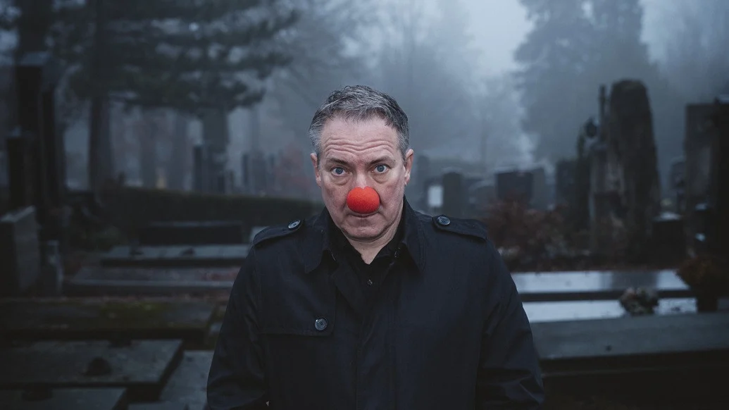 Man in a dark coat with a red clown nose standing in a misty cemetery.