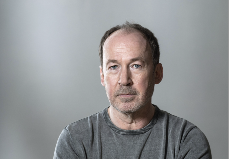 Portrait of a middle-aged man with short brown hair and stubble, wearing a gray crewneck shirt, against a neutral gray background.
