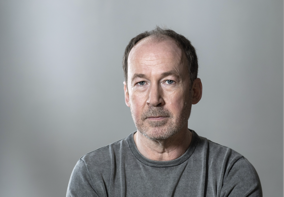 Portrait of a middle-aged man with short brown hair and stubble, wearing a gray crewneck shirt, against a neutral gray background.