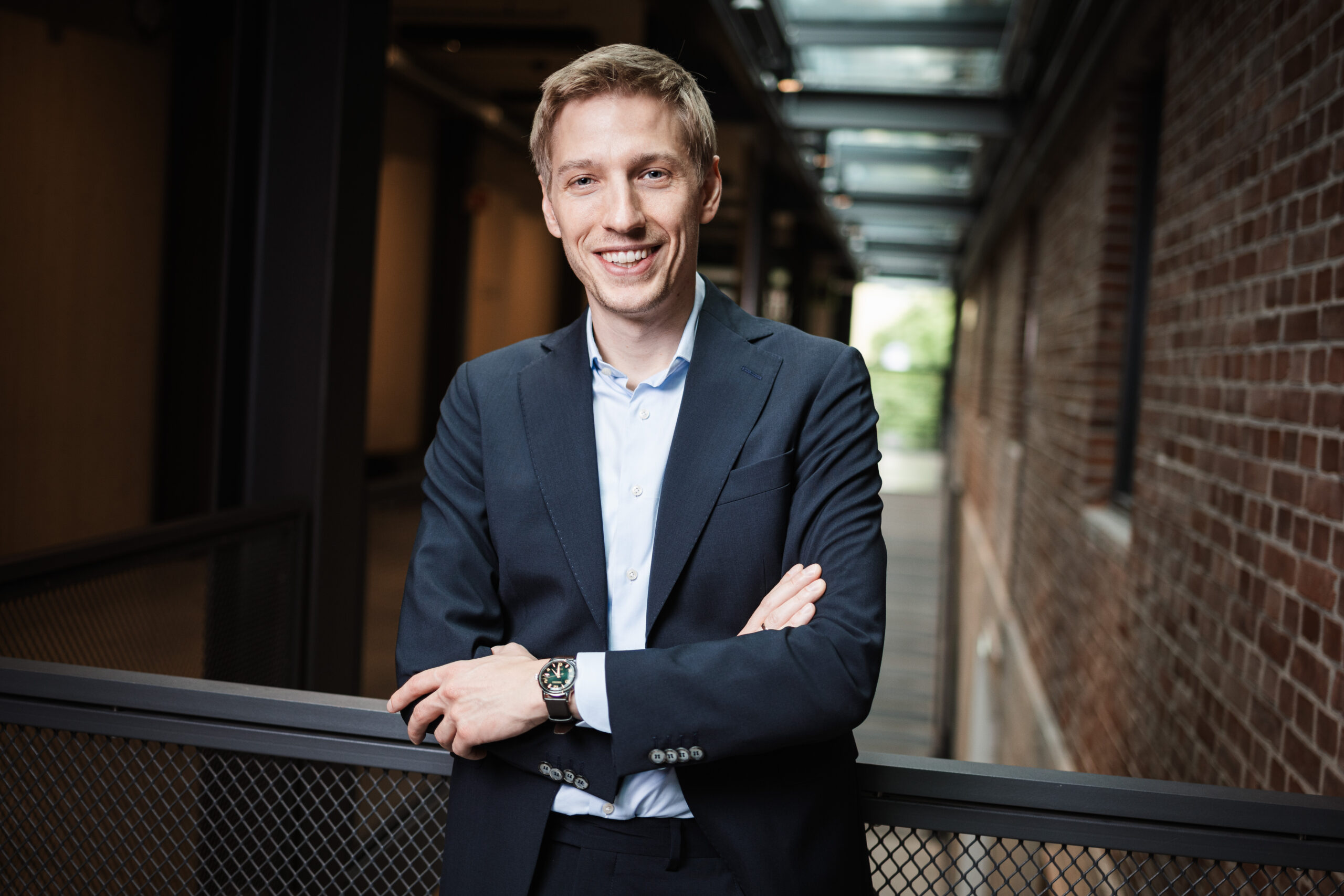 Smiling man in a navy suit leaning on a railing in a brick-walled corridor outdoor setting.