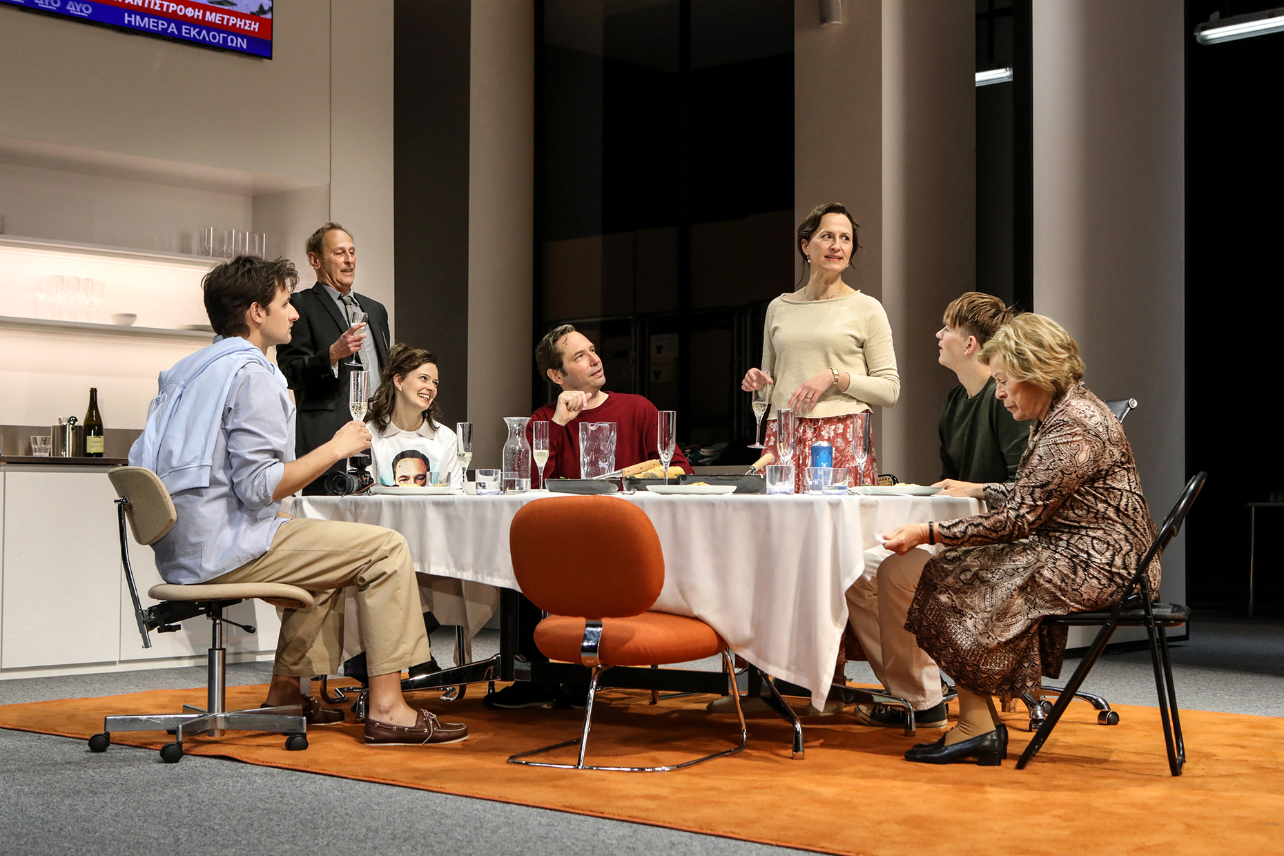 A group of adults sits around a dining table toasting with champagne in a modern office kitchen setting, listening to a woman who stands and speaks.