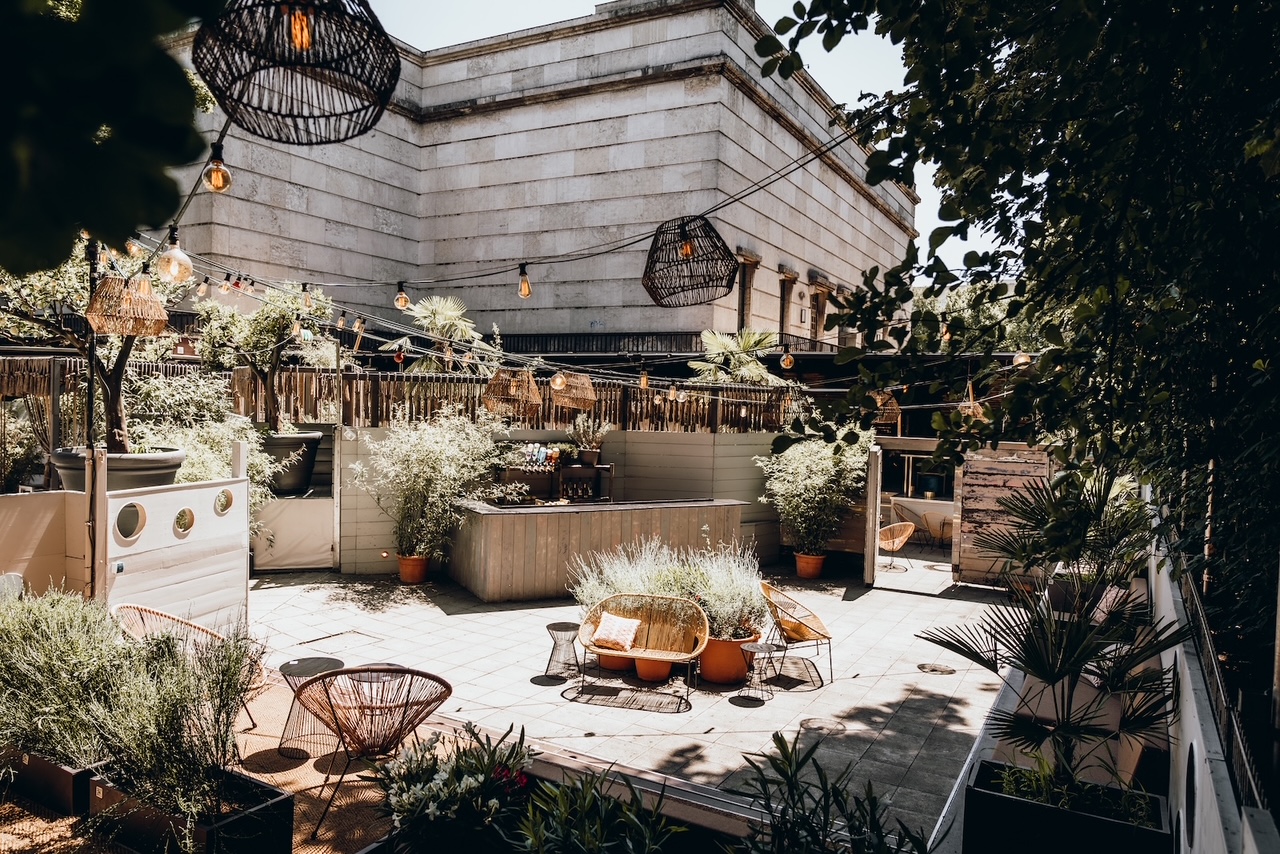 Sunlit rooftop terrace with wicker chairs, low tables, and string lights among potted plants and greenery.