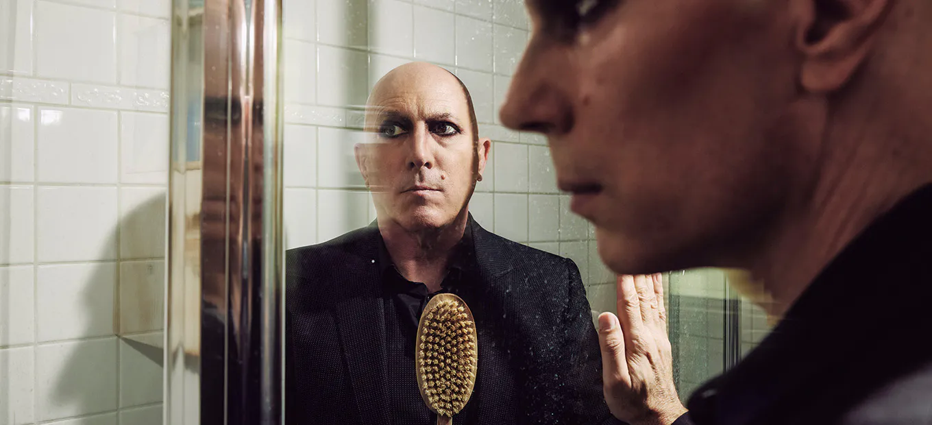 Man in a dark suit seen through a glass shower door, staring intently at his reflection while a wooden bristle brush rests on his chest.