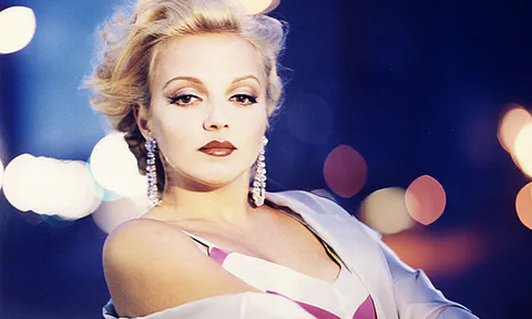 Portrait of a glamorous woman with blonde hair, bold makeup, and dangling earrings against a city lights backdrop.