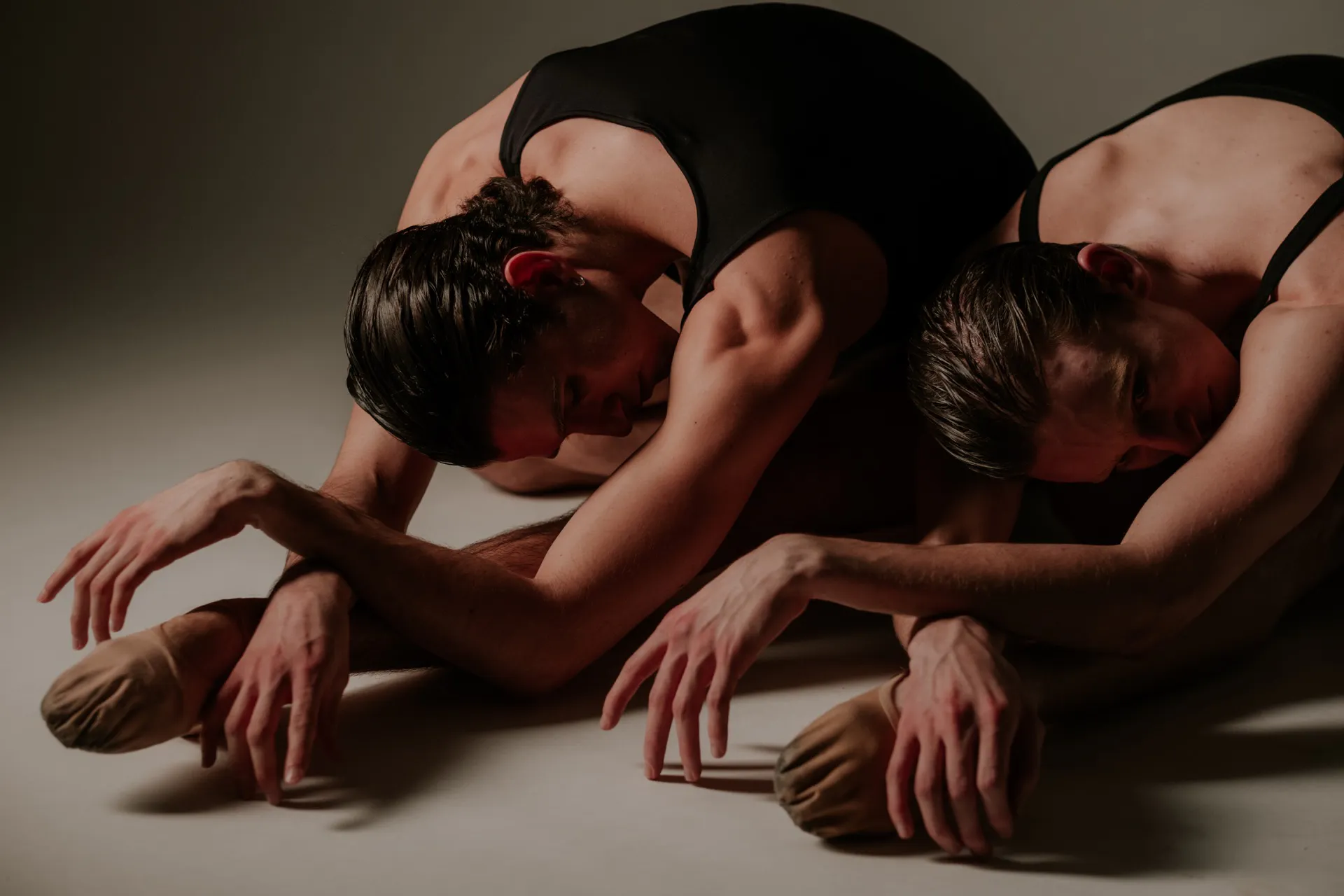 Three dancers in black leotards bend forward on the floor, arms extended in a synchronized floor pose in a dim studio light.