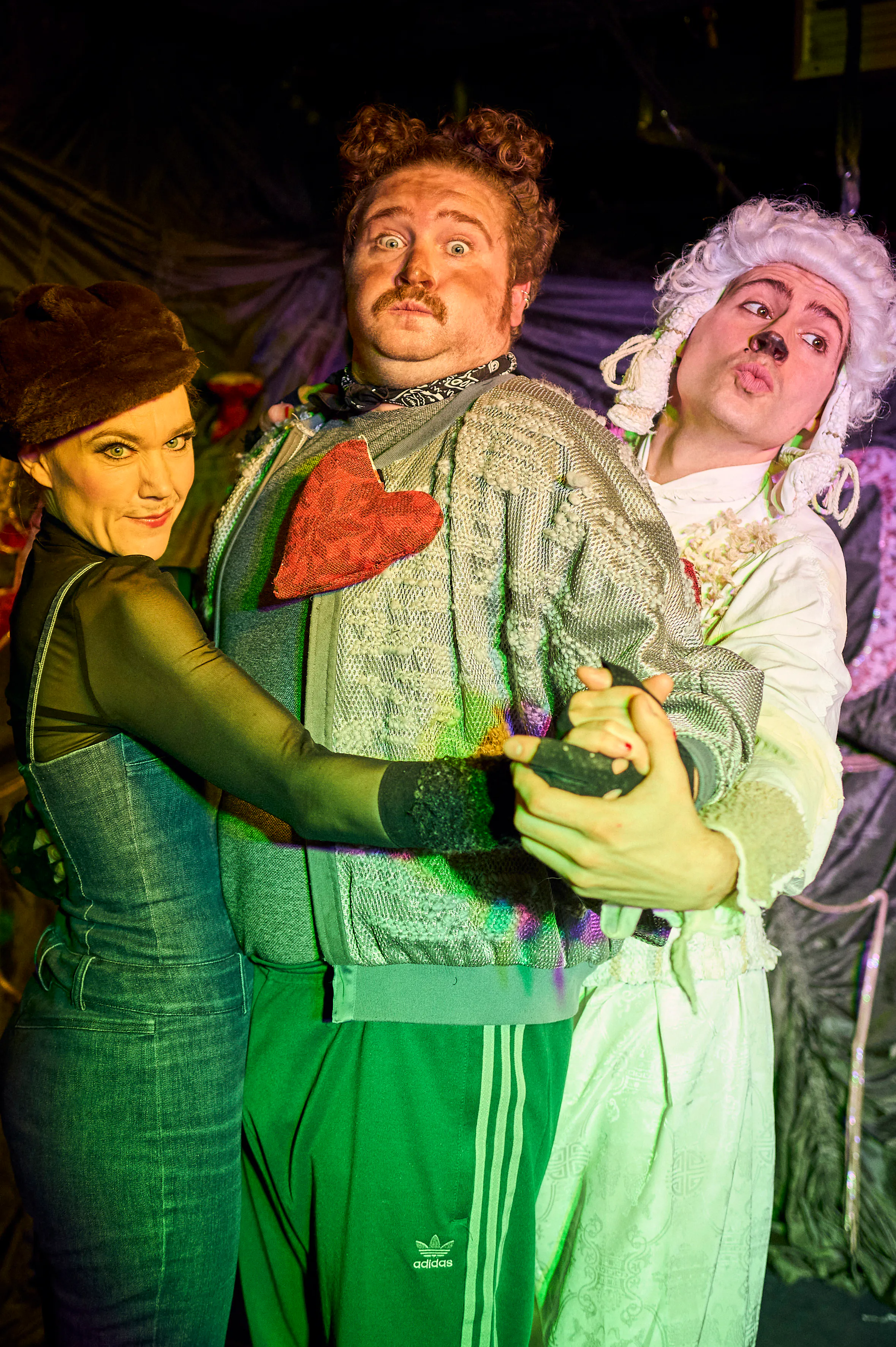 Three costumed performers pose closely, embracing; center man with mustache in metallic jacket and red heart on chest, flanked by a woman in brown hat and a white-wigged performer in pale makeup.
