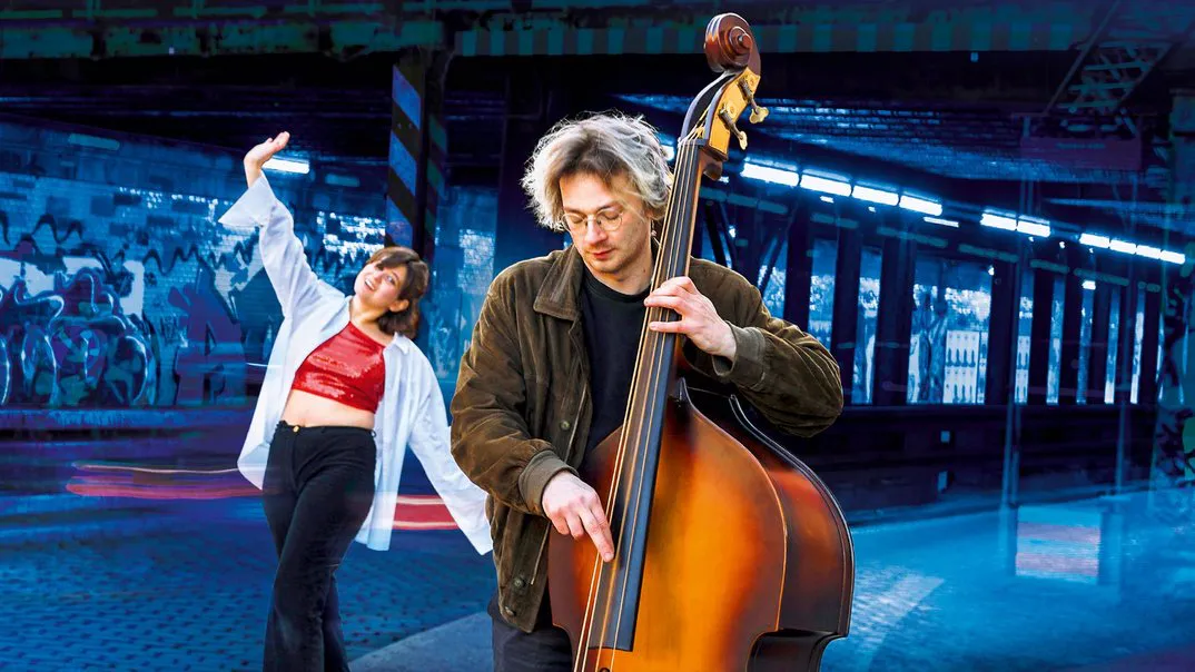 Musician playing a double bass in a graffiti-covered subway station while a woman dancer moves energetically in the background.