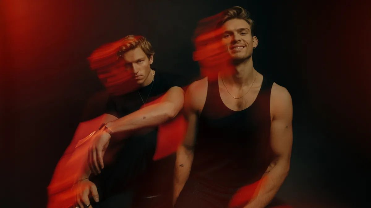 Two young men pose in a dark studio with red light streaks creating a motion blur around them on a moody background.