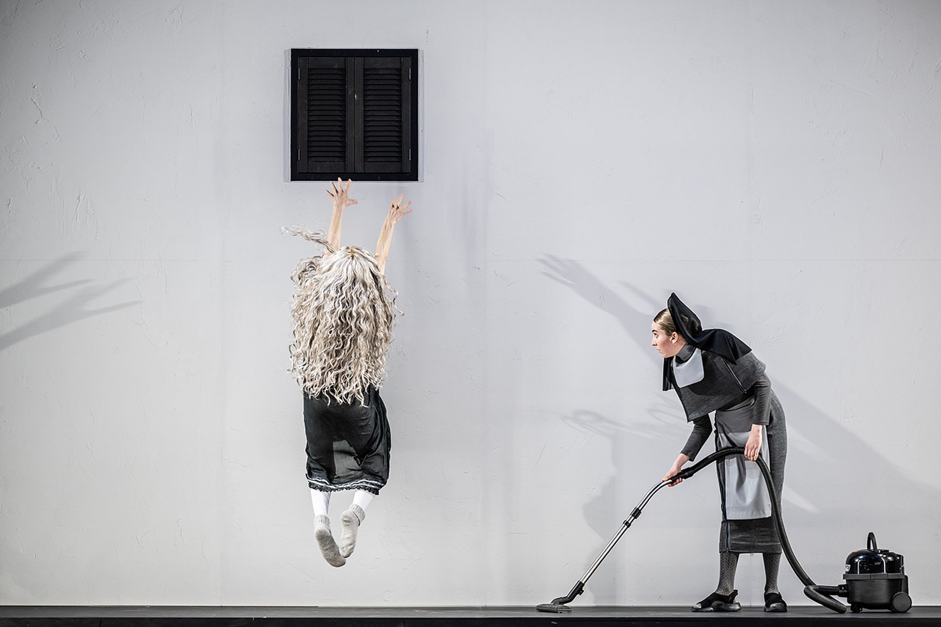 Stage scene with a dancer leaping near a gray wall, wearing a curly blonde wig; a second performer in gray costume uses a vacuum cleaner nearby.